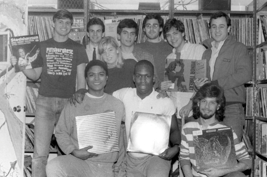 Black and white photo of WSRN student workers in the station, surrounded by records. Some of them hold records, by artists such as Stockhausen, Minor Threat, and Rhinoceros. Other album covers are unreadable due to the lighting.