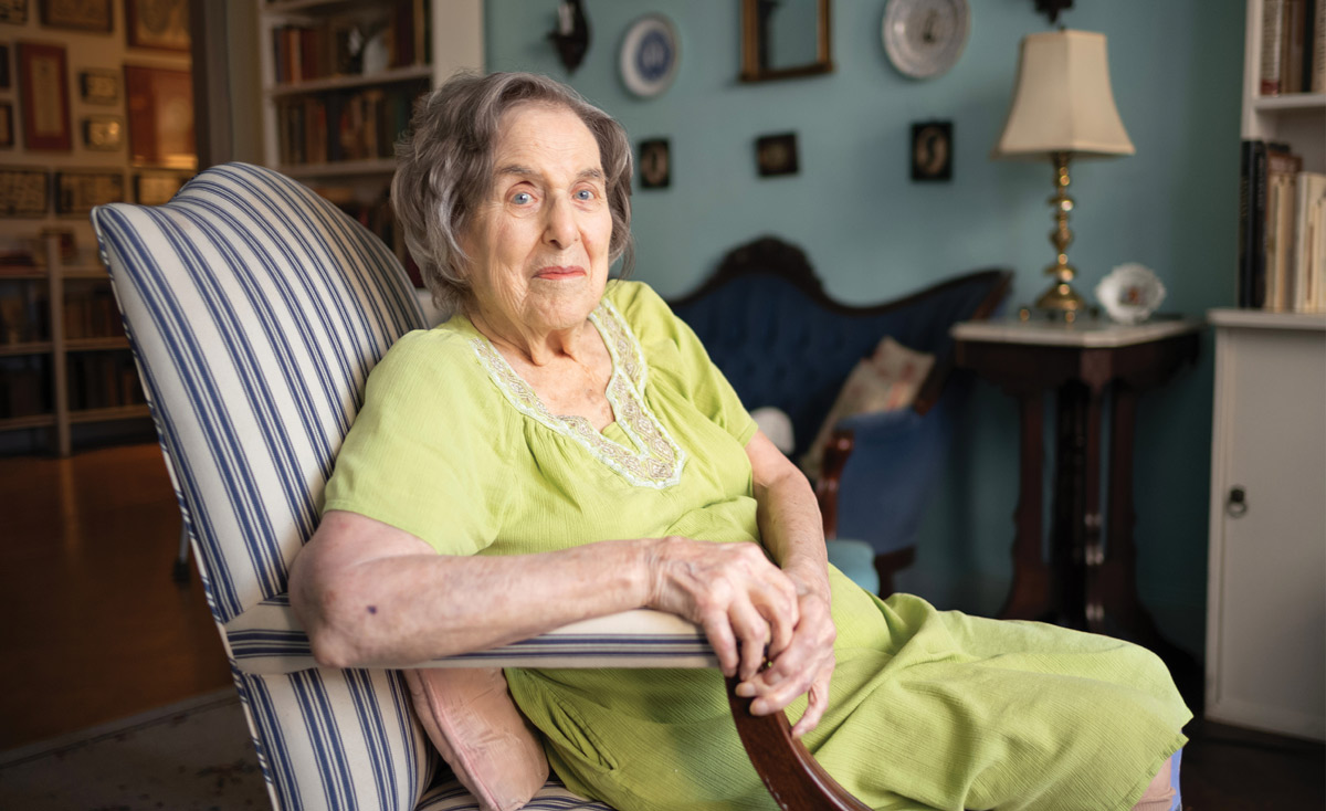 Lucy Seligman gazes at the camera from her blue-and-white striped armchair. She has blue eyes and wears a lime green dress with an embroidered collar.