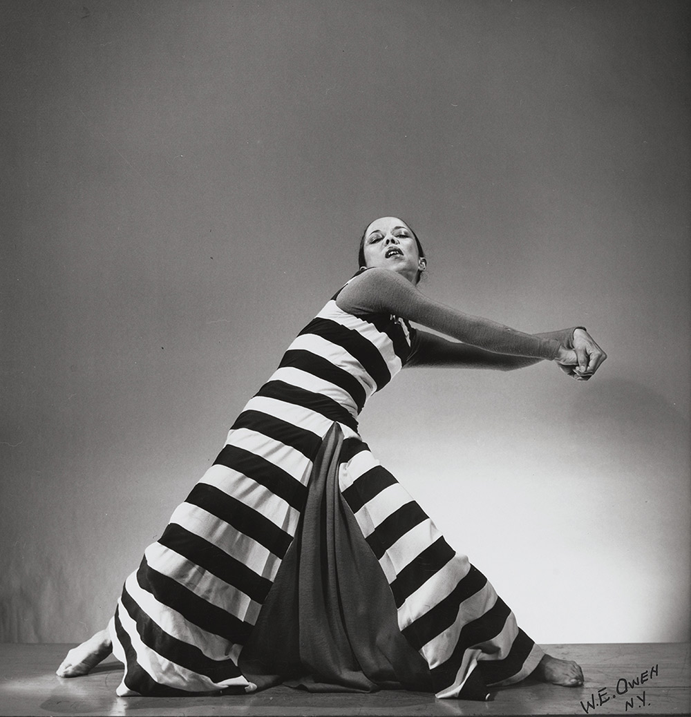 Janet Collins in "Chain Gang." In this black and white photo, she wears a long striped dress and clasps her hands to the right side of her body.