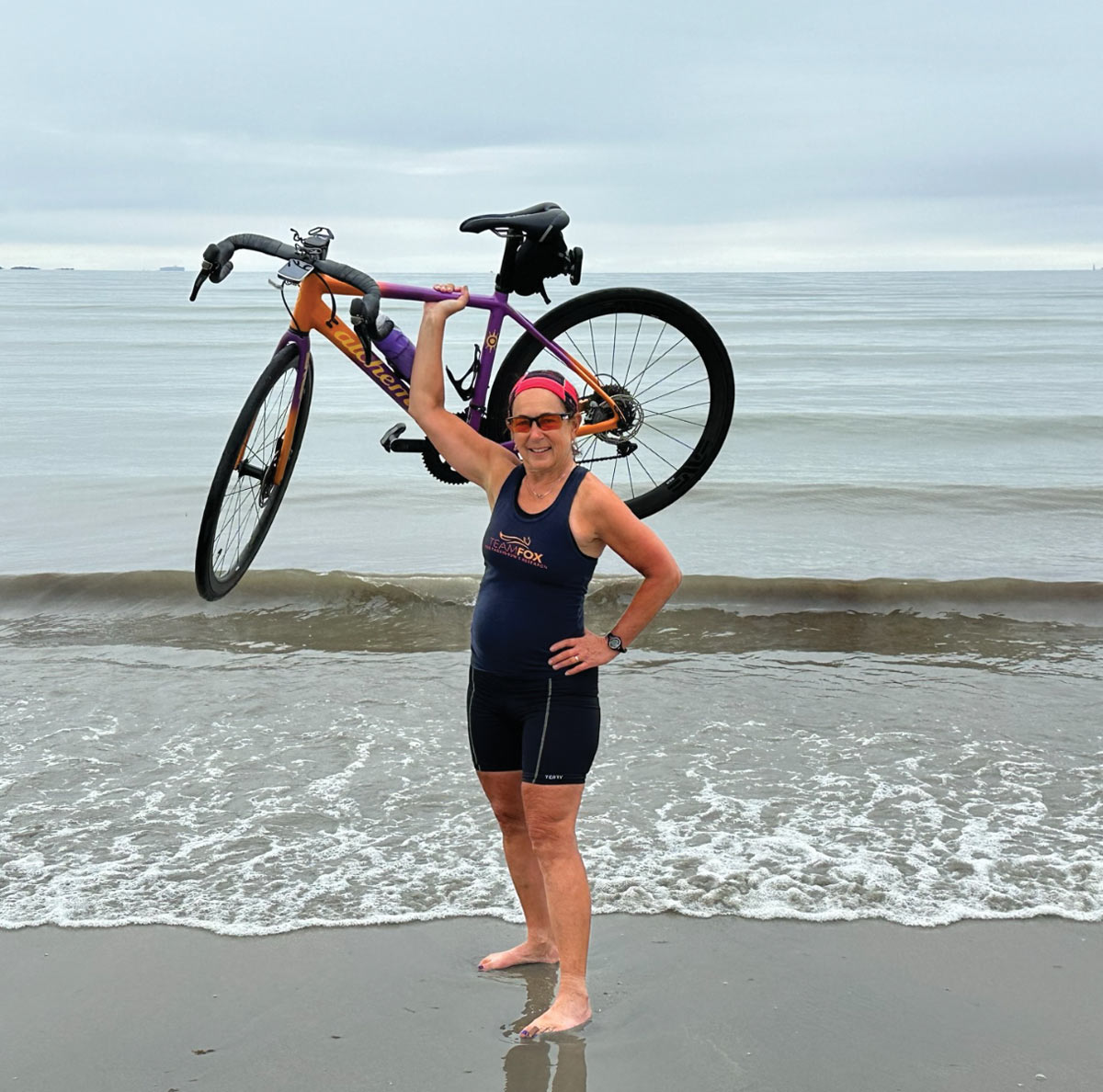 Barbara Harris stands in shallow waves on a beach, holding her bike over her head with one hand.