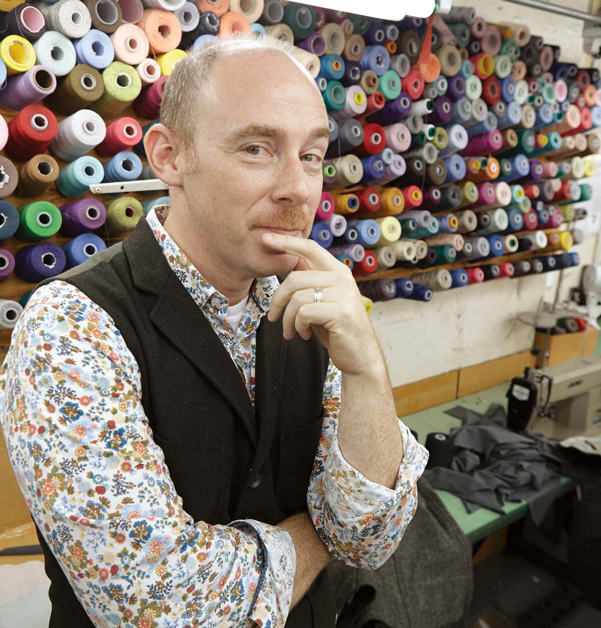 Alexander Joseph stands by a sewing machine, with spools of colorful thread behind him.