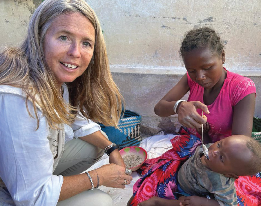 In an outdoor camp providing humanitarian aid, Allison Oman Lawi looks toward the camera in a white shirt and is kneeling beside a woman with a serious expression in pink shirt who is holding a small child and feeding the child with a spoon.