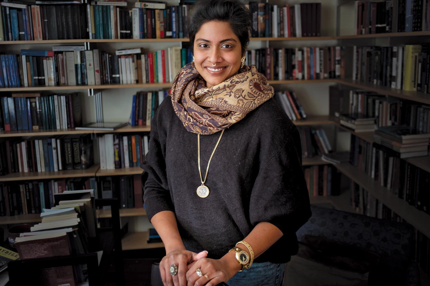 Headshot of Sabeen Ahmed in her office, surrounded by books. She wears a black shirt, beige patterned scarf and a bright gold medallion necklace.