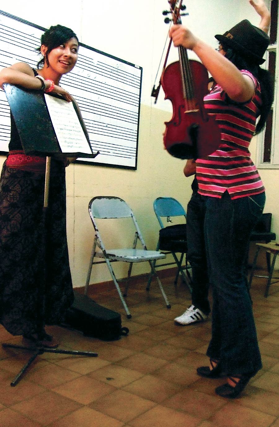 Stephanie Hsu (left) turns pages of sheet music for a student (right) who holds a violin and stands on their tiptoes.