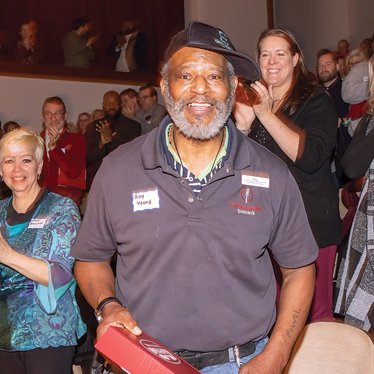 Roy Wood smiles, holding his award and facing the camera, while his colleagues applaud him in the background.