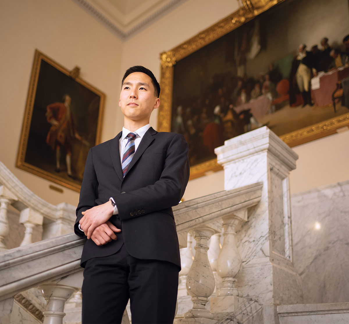 Jason Heo poses on the staircase of the Maryland State House. He is wearing a suit and tie and has his hands crossed in front of him.