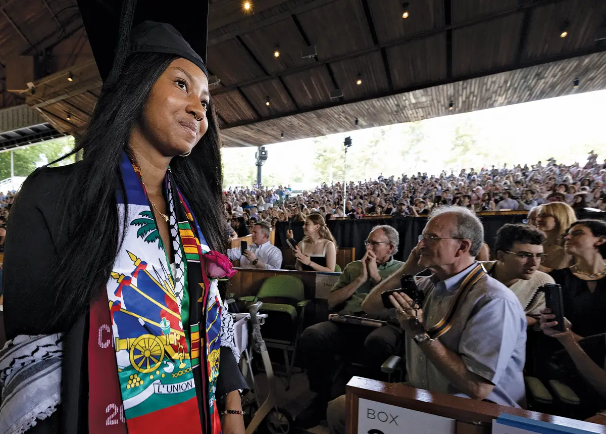 Student crosses the graduation stage, proudly wearing the Haitian flag, while crowd applauds.