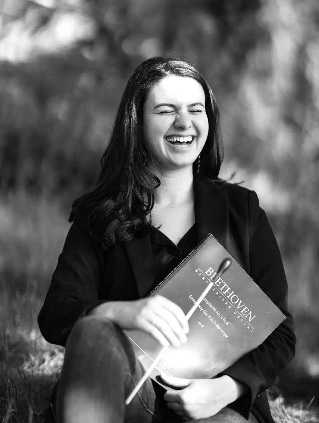 Black and white photo of Shira Samuels-Shragg, smiling, holding a baton and a book of Beethoven's music.