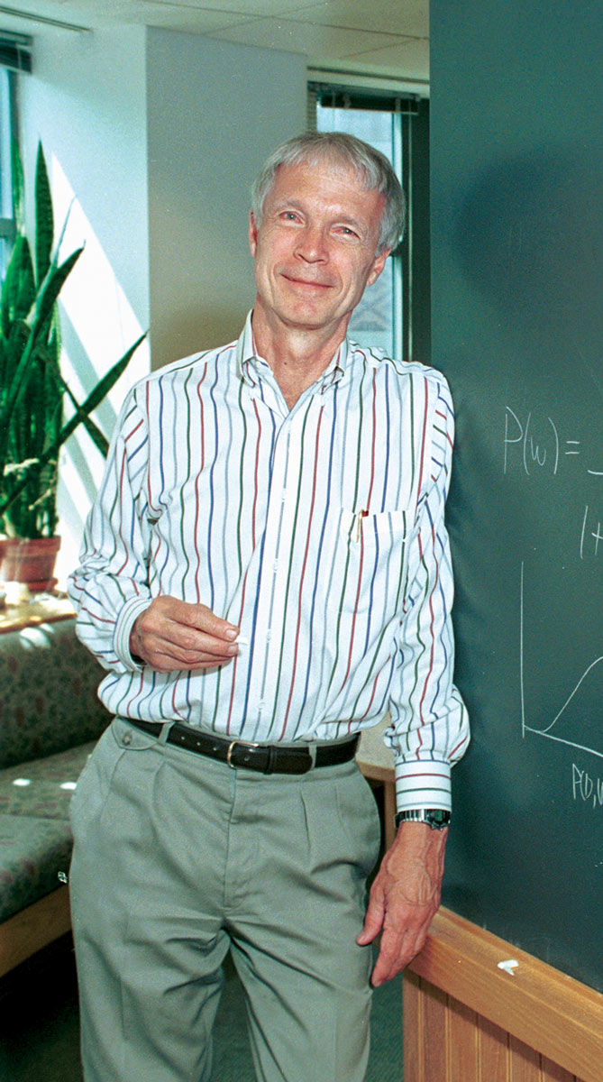 John Hopfield leans against a blackboard holding a piece of chalk.
