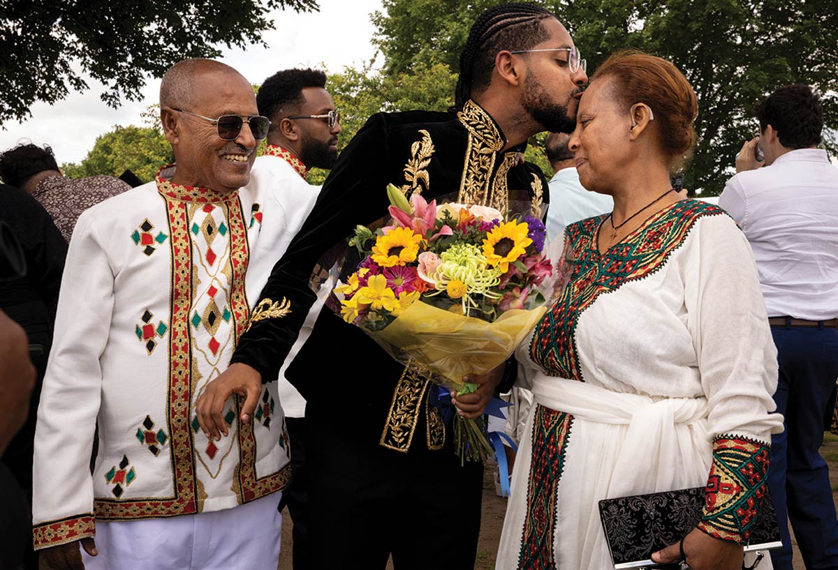 A student kisses his mom’s forehead while his dad watches on, smiling.