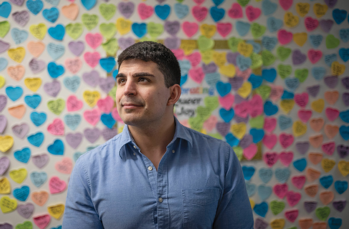Lucas Zullo looks off to the left, wearing a denim blue button-down. Behind him on the wall are hundreds of colorful heart-shaped sticky notes.