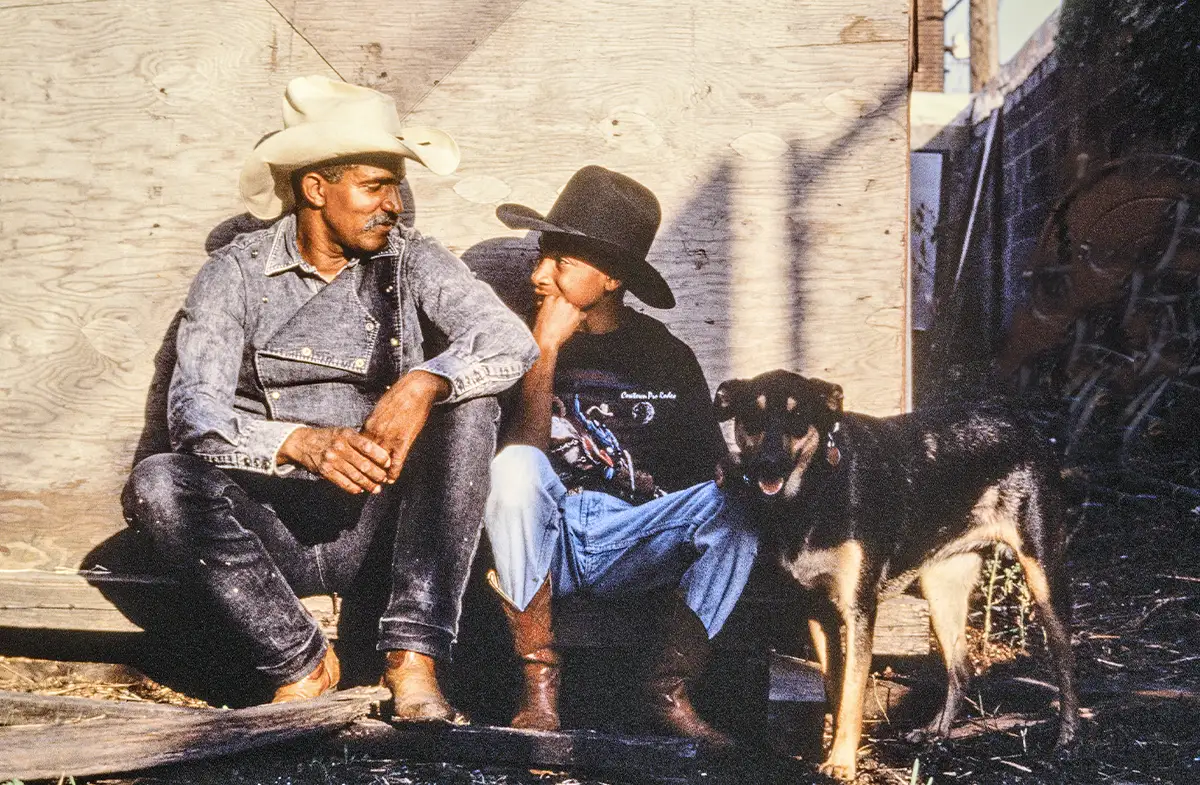 An older man and a younger man sit, looking at each other. Both wear cowboy hats. There’s a dog on their right.