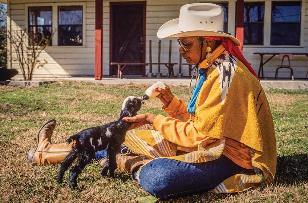 A woman in a cowboy hat feeds a baby goat milk from a bottle.