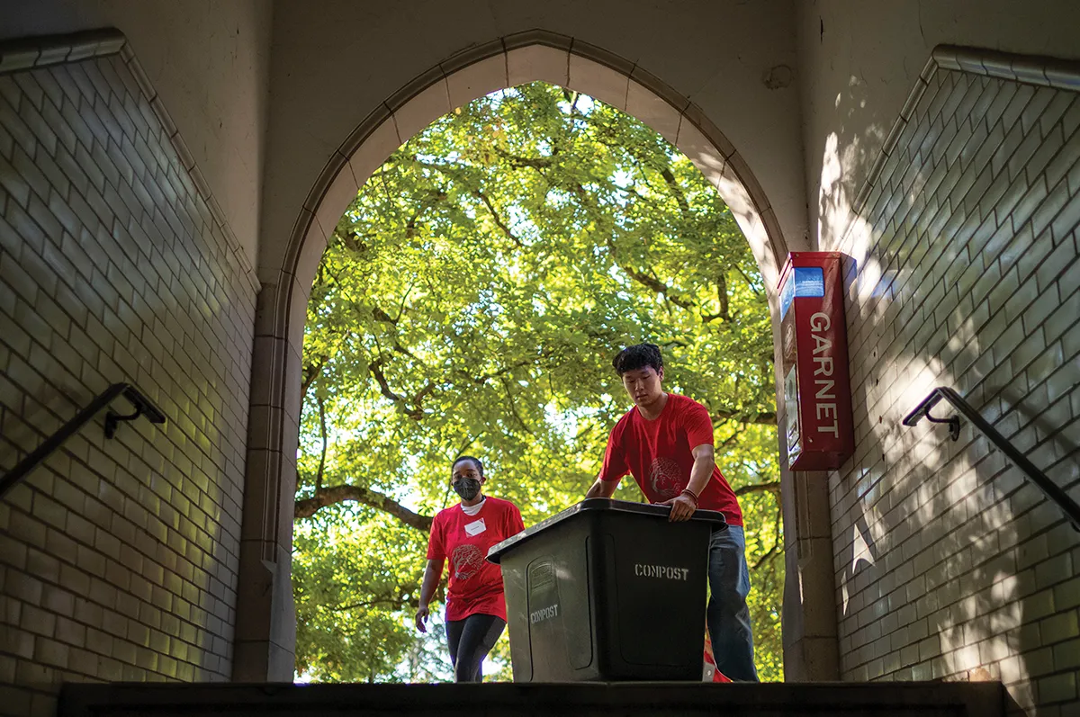 Two students in red shirts, framed by a stone archway, move a large compost bin