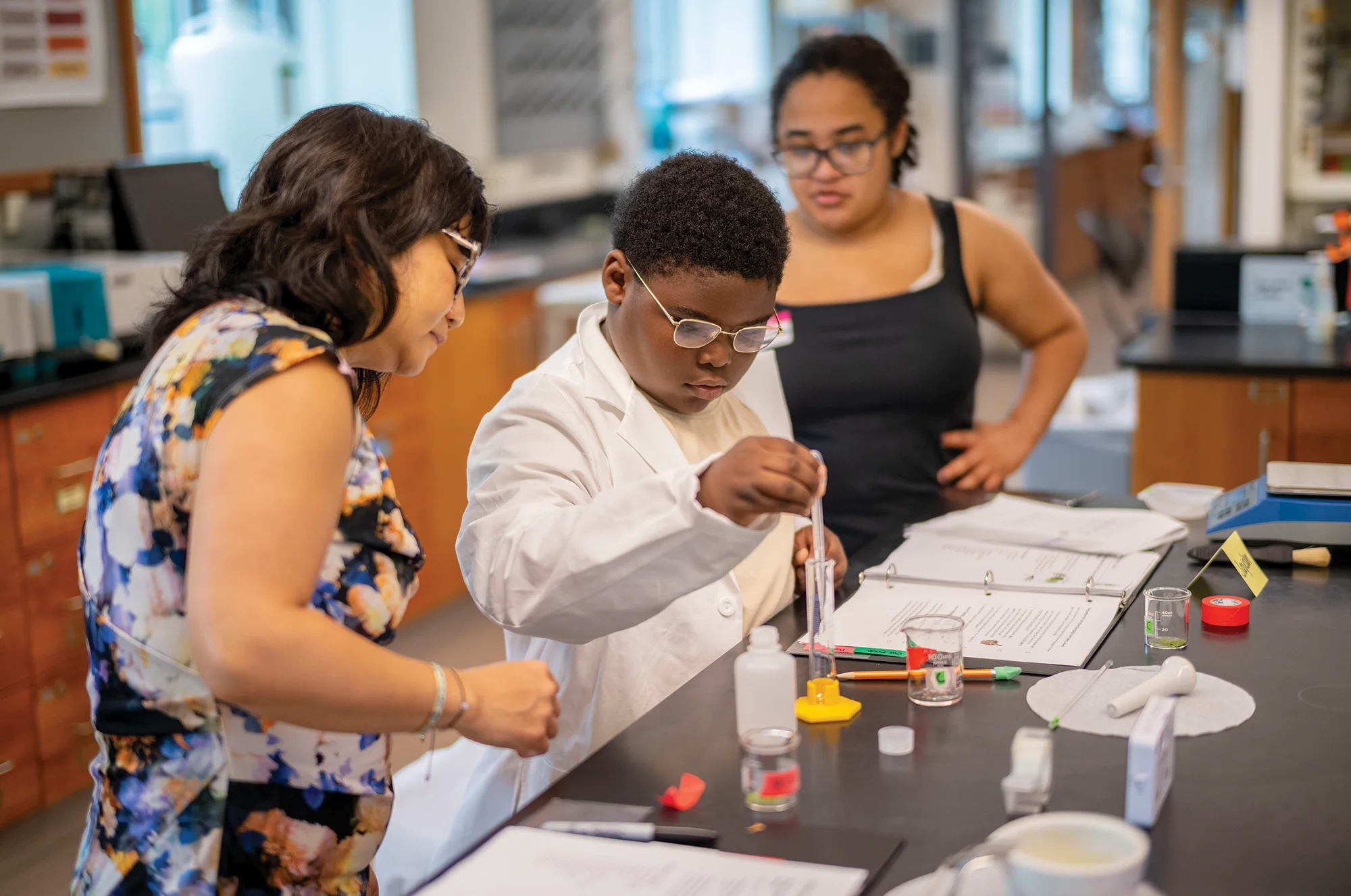 A young student in a lab coat pours liquid into a beaker from a pipette.
