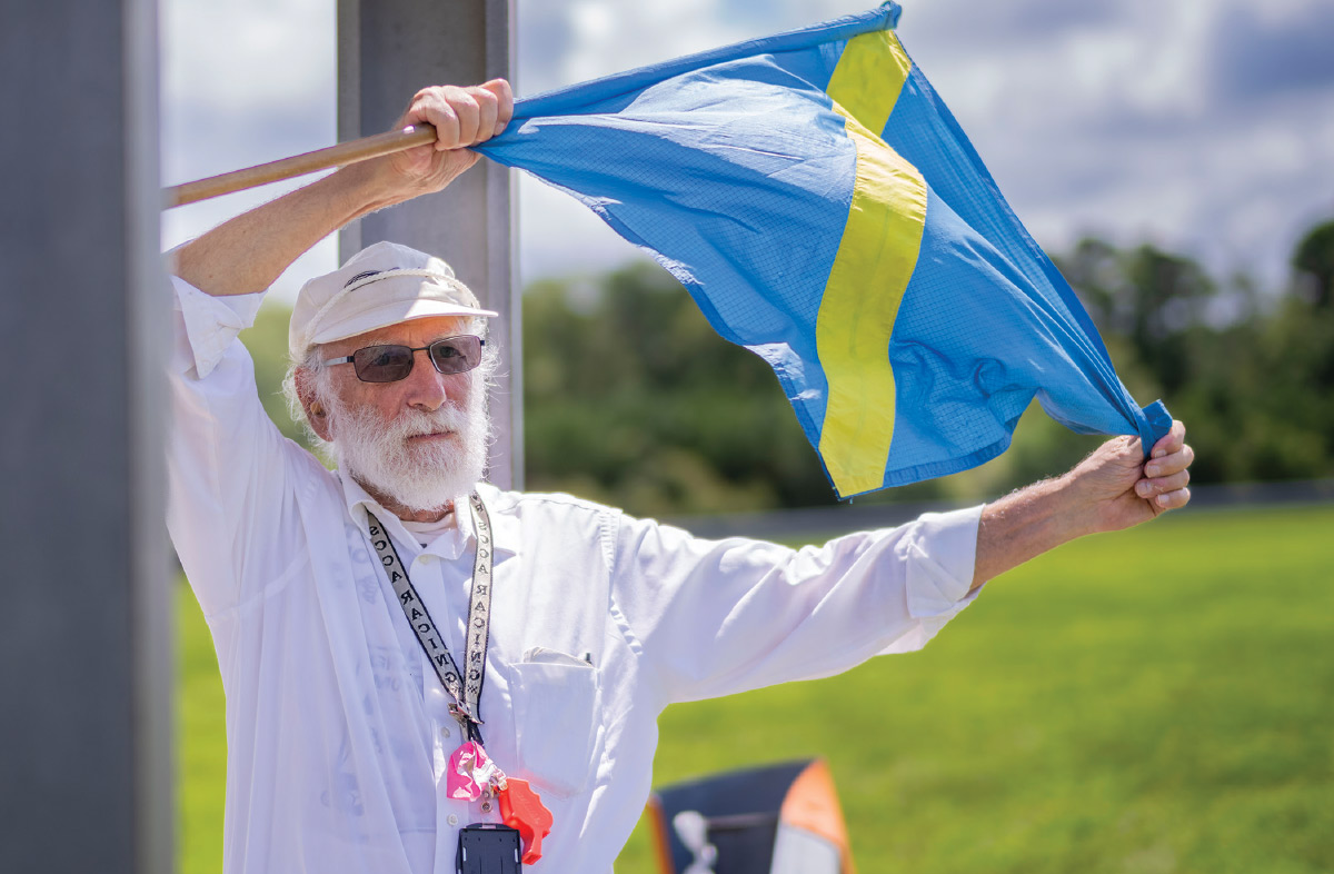 Peter Meyer ’65 waves a blue flag with a yellow stripe crossing it diagonally.