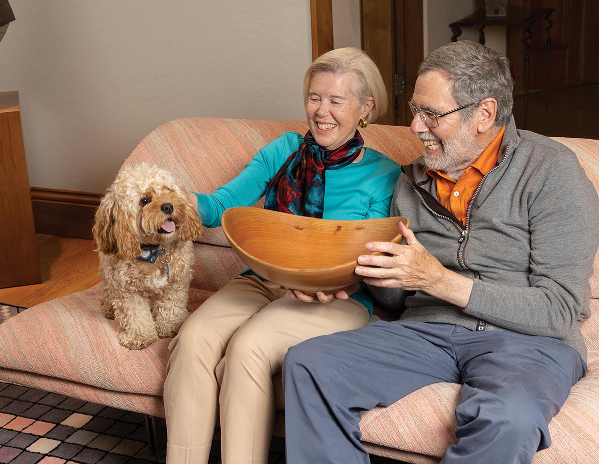 Roy holds the wooden bowl while Linda pets the family dog.