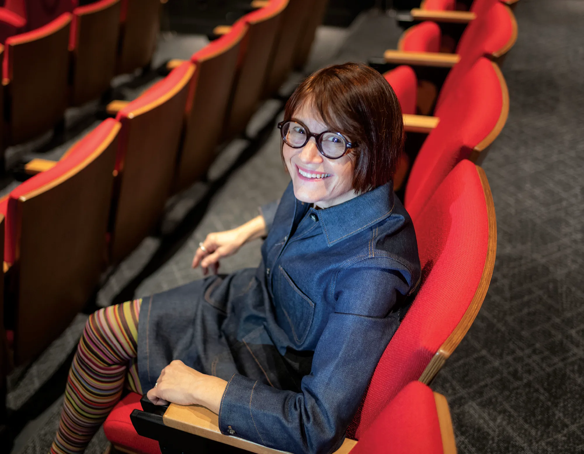 Patricia White smiles at the camera from the new screening room in Martin Hall. She wears a denim dress, striped leggings, and black glasses, and sits in a bright red seat.