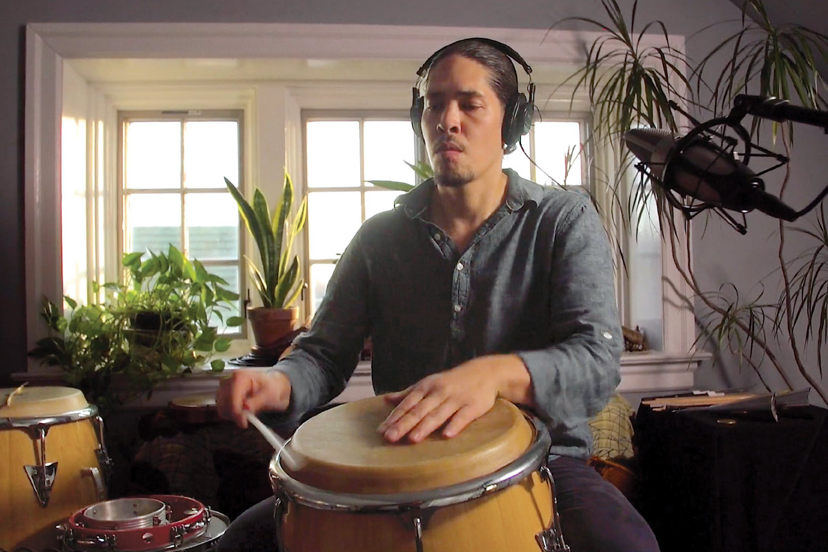 Alex Shaw '00 records a percussion track in front of a window filled with houseplants.