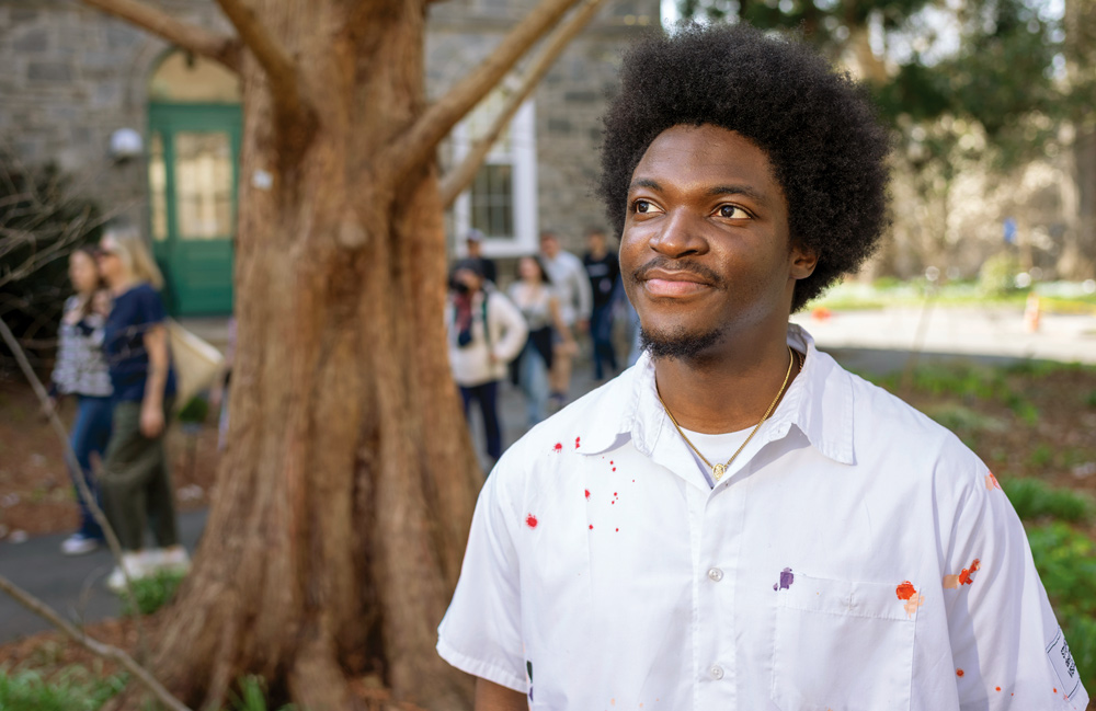 Steven Mukum '26 looks up to the left and smiles. He wears a white button-down shirt with splashes of different colors of paint on it.