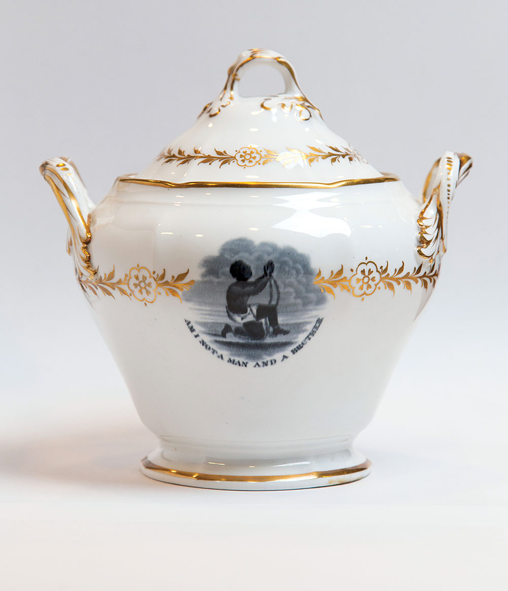 A white sugar bowl with gold trim and a drawing of a Black man chains with the text "Am I not a man and a brother?" underneath the image.
