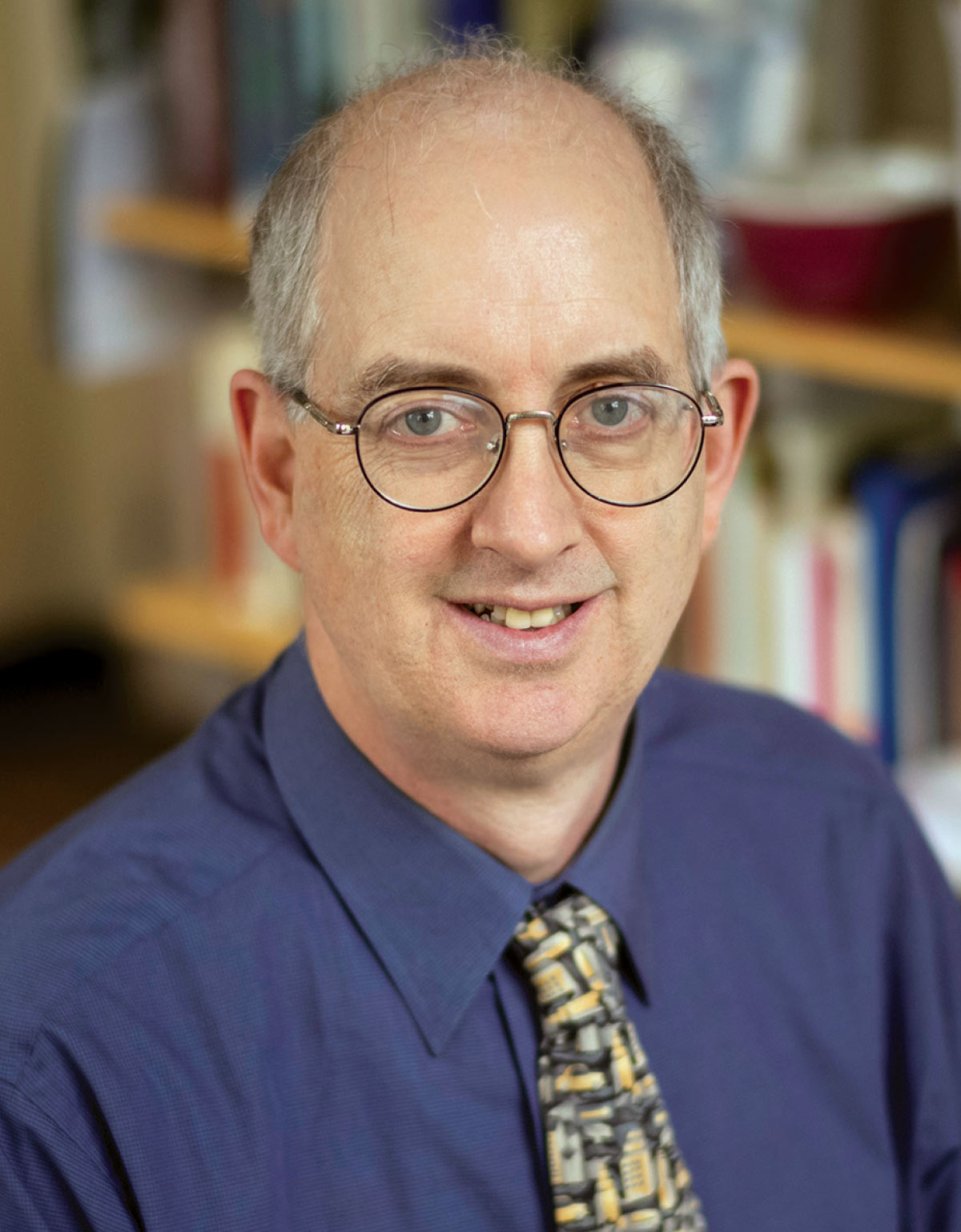 Head shot of Alan Baker wearing glasses, a blue button-down shirt, and a tie with a yellow pattern.