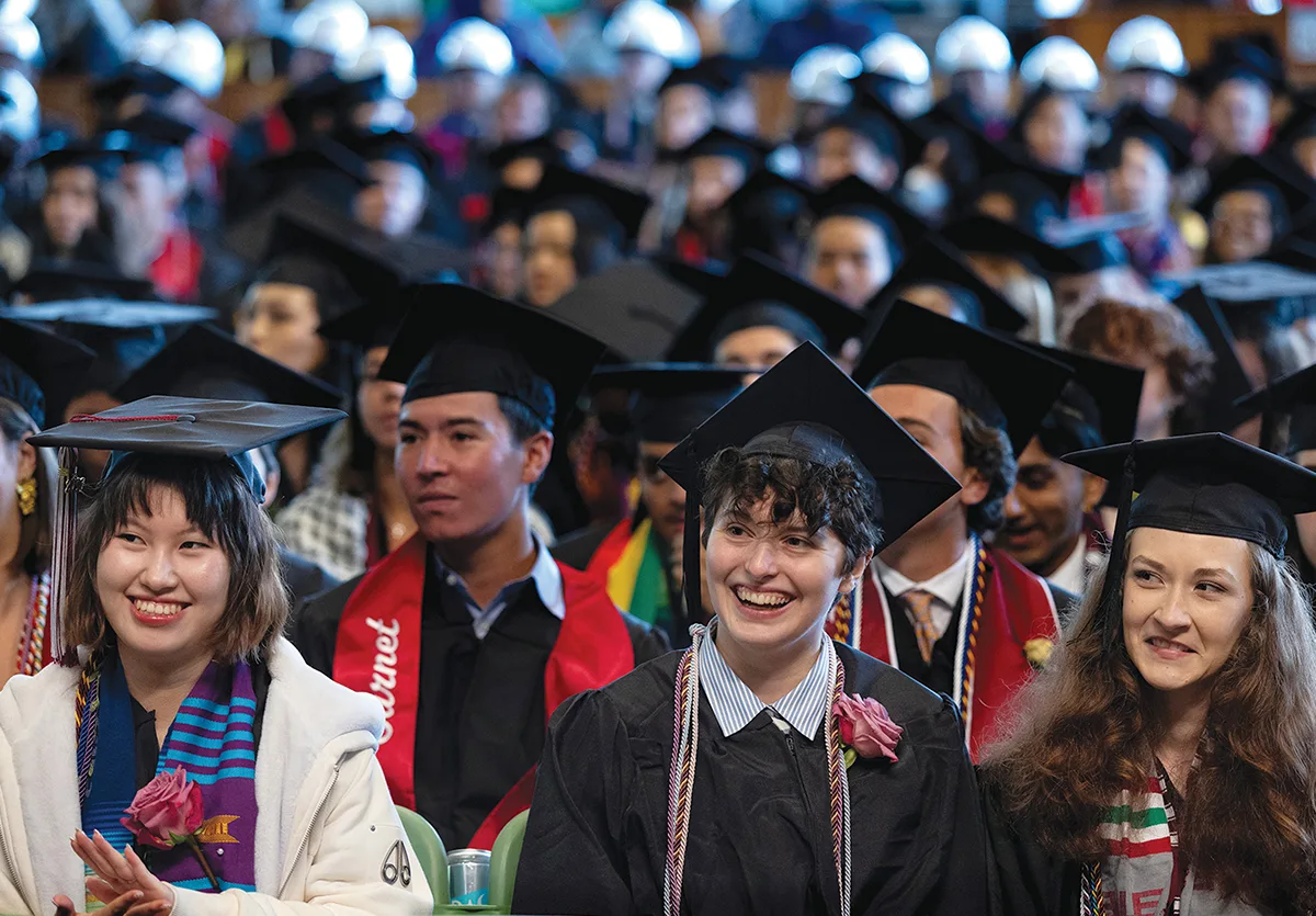 Students in caps and gowns smile and clap for their fellow graduates.