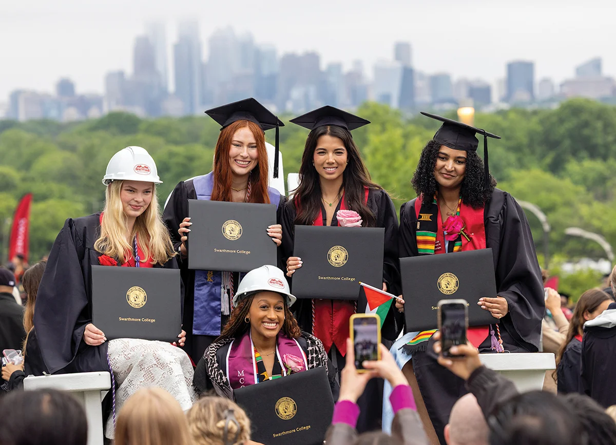 Five female students of different races smile and pose for a photo with their newly earned degrees. Three wear the traditional cap and gown and two (engineering majors) wear hard hats with their gowns.