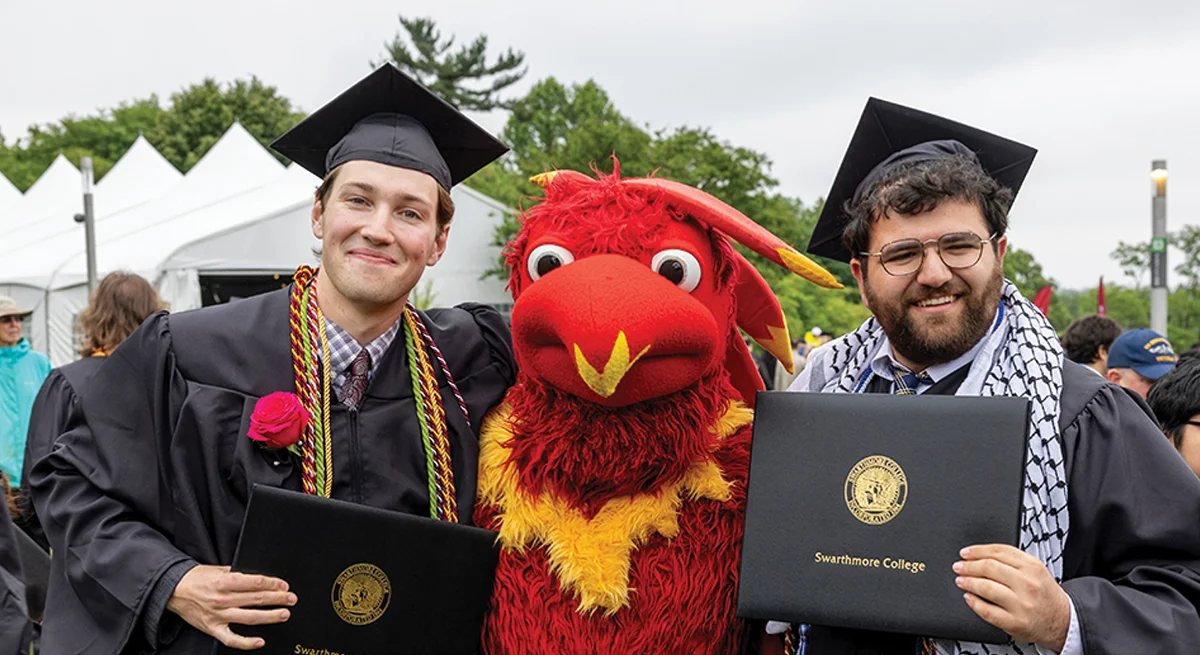Two students pose, with Phineas the Phoenix and their newly earned diplomas, in their caps and gowns.