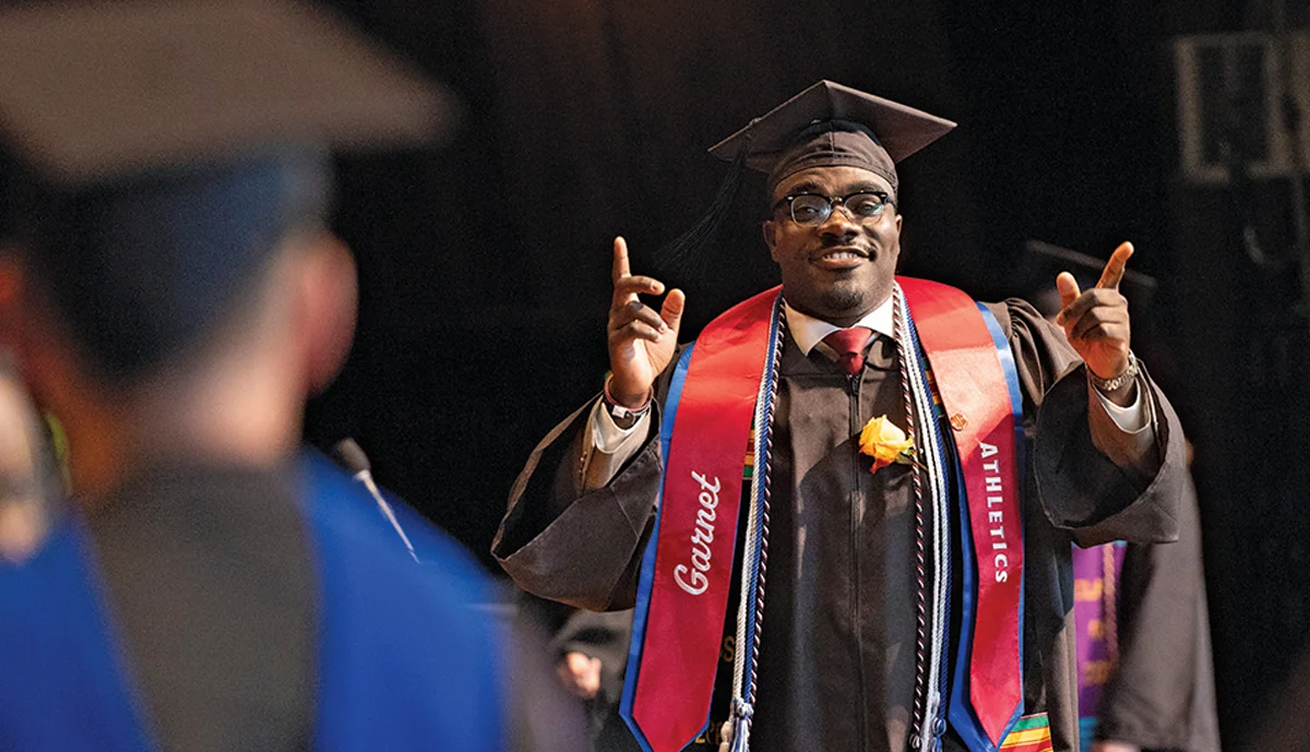 A students smiles and points while crossing the stage wearing a cap, gown, and Garnet Athletics sash.